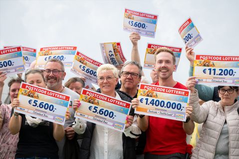 A group of people happily holding large checks from the Postcode Lottery, with amounts ranging from €5,600 to €1,400,000.
