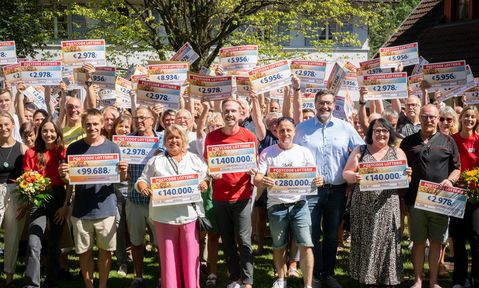 A group of people outdoors holding large checks with various amounts, smiling and celebrating under a tree.