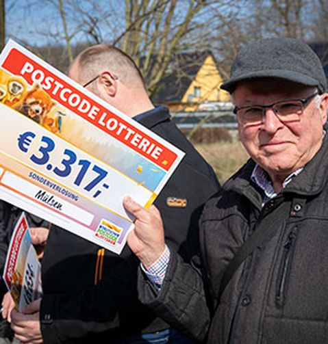Ein Mann hält einen Postcode-Lotterie-Scheck über 3.317 € und steht im Freien mit weiteren Personen.
