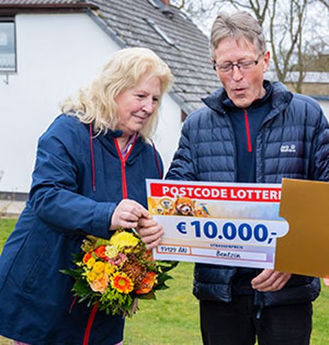 Zwei Personen halten einen großen Scheck über 10.000 € von der Postcode Lotterie. Eine von ihnen trägt einen Blumenstrauß. Sie stehen im Freien in der Nähe eines Hauses.