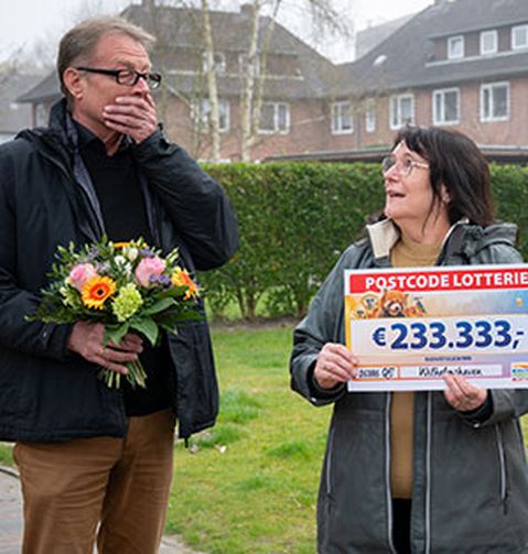 Ein überraschter Mann hält einen Blumenstrauß, während eine Frau in einem Wohngebiet einen Postcode-Lotterie-Scheck über 233.333 € präsentiert.