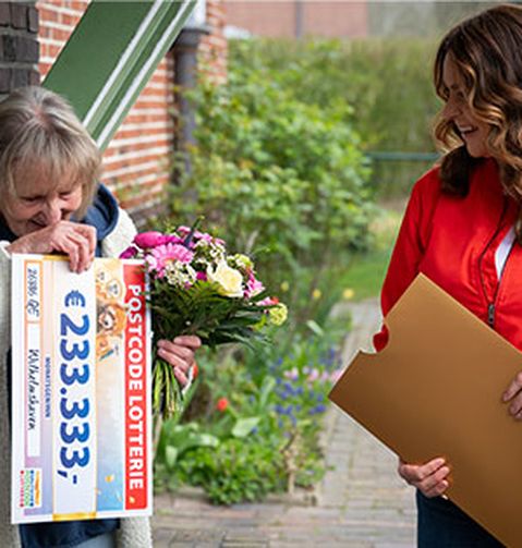 Eine fröhliche Frau hält einen Blumenstrauß und einen großen Lotterie-Scheck und lächelt eine andere Frau in roter Jacke an, die einen Umschlag in der Hand hält.