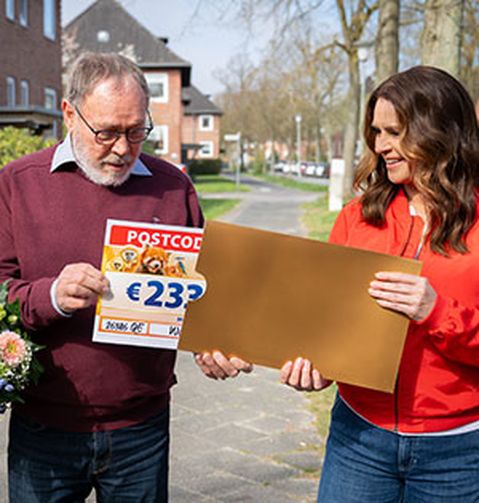 Eine lächelnde Frau überreicht einem älteren Paar auf einer sonnigen Straße einen Umschlag. Das Paar hält einen Lotterie-Scheck und Blumen in den Händen.