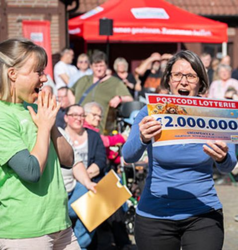 Zwei Frauen feiern im Freien, eine von ihnen hält einen großen Scheck über 2.000.000 € von einer Lotterie. Sie sind von einer jubelnden Menschenmenge umgeben.