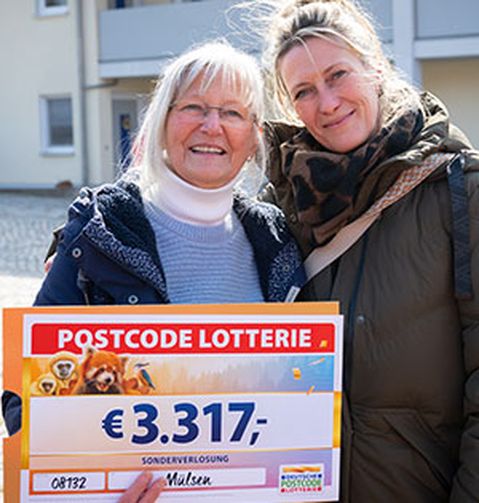 Zwei Frauen lächeln und halten einen großen Lotterie-Scheck über 3.317 €. Sie stehen im Freien in einem Wohngebiet.