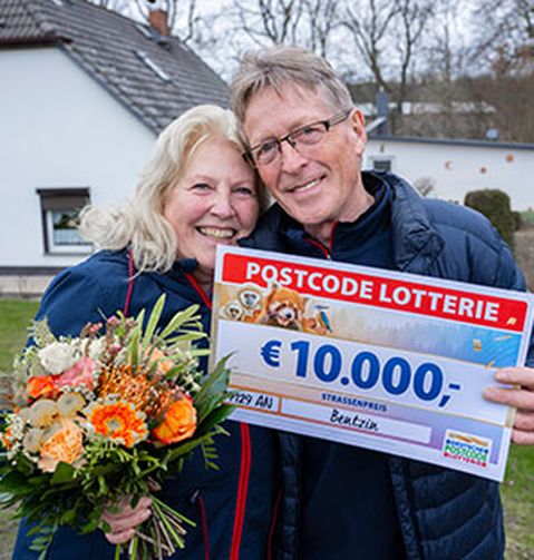 Ein lächelndes Paar hält einen großen Lotterie-Scheck über 10.000 € und einen Blumenstrauß in den Händen. Sie stehen im Freien in der Nähe eines Hauses.