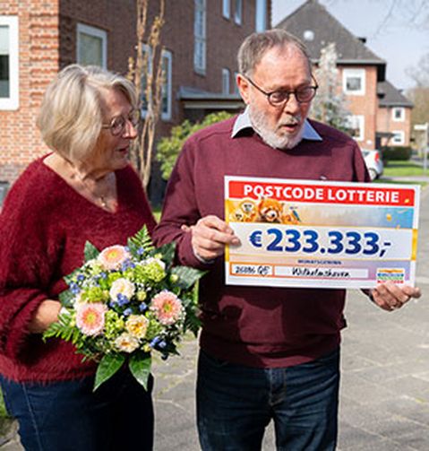 Drei Personen stehen im Freien: Eine hält einen großen Lotterie-Scheck über 233.333 €, eine andere einen Blumenstrauß, und eine Frau mit einem Umschlag lächelt.
