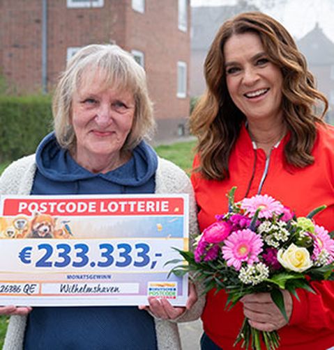 Zwei Frauen lächeln, eine von ihnen hält einen „Postcode Lotterie“-Scheck über 233.333 €, die andere einen Blumenstrauß.