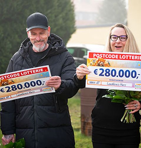 Zwei Personen lächeln und halten große Schecks über 280.000 € von der Postcode-Lotterie. Sie stehen im Freien, im Hintergrund ist viel Grün zu sehen.