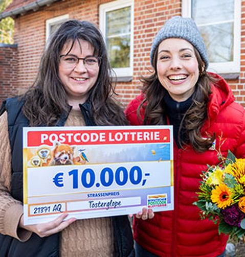 Zwei Frauen stehen vor einem Haus. Eine hält einen großen Umschlag, die andere ein Schild mit der Aufschrift „10.000 €“ und „Postcode“.