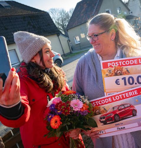 Zwei Frauen lächeln. Eine von ihnen hält einen Blumenstrauß und Lotterie-Schecks, die andere macht ein Selfie. Sie befinden sich im Freien in einem Wohngebiet.