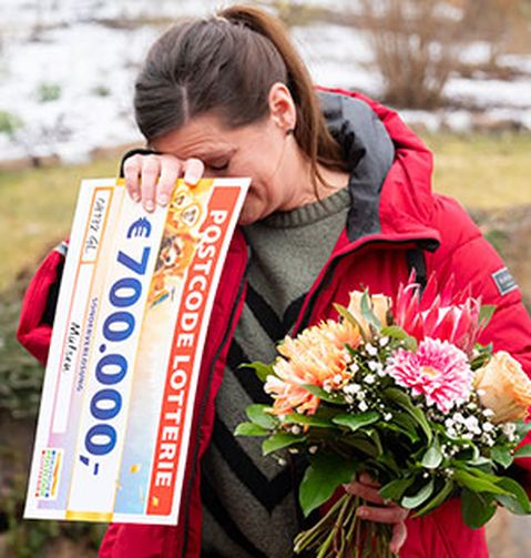 Eine Frau in roter Jacke hält einen Blumenstrauß und einen großen Lotterie-Scheck über 700.000 €. Sie wirkt gerührt und bedeckt mit einer Hand ihr Gesicht.