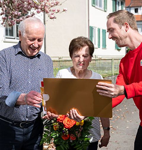Drei Personen stehen im Freien, eine von ihnen hält einen Blumenstrauß, eine andere einen großen goldenen Umschlag. Sie lächeln und scheinen in ein Gespräch vertieft zu sein.