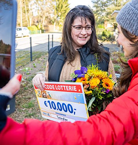 Eine lächelnde Frau hält einen Lotterie-Scheck über 10.000 € und einen Blumenstrauß in den Händen, umgeben von weiteren Personen im Freien.