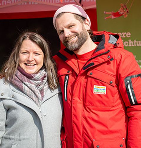 Ein Mann und eine Frau lächeln im Freien und tragen Winterjacken. Der Mann trägt eine rote Jacke und eine rosafarbene Mütze, die Frau einen grauen Schal und einen Mantel.