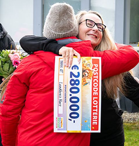 Zwei Personen umarmen sich freudig, eine von ihnen hält einen großen Lotterie-Scheck über 280.000 € von der Postcode Lotterie. Im Hintergrund sind Blumen zu sehen.