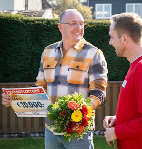 Ein Mann hält einen Scheck über 10.000 € und einen Blumenstrauß in den Händen und lächelt einen anderen Mann in einem Garten an.
