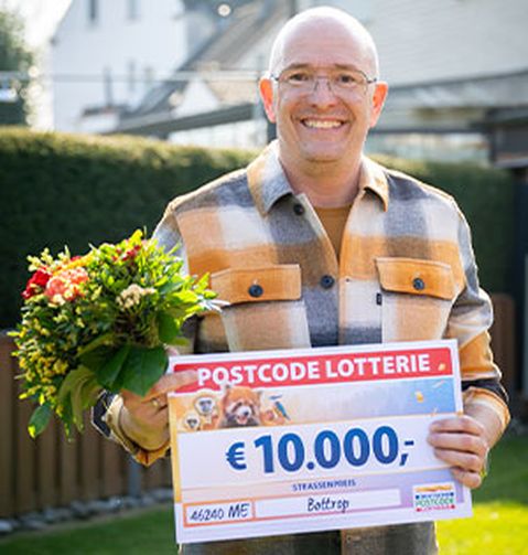 Ein Mann lächelt, hält einen Blumenstrauß und einen großen Scheck über 10.000 € von der Postcode Lotterie. Er steht im Freien in einem Garten.