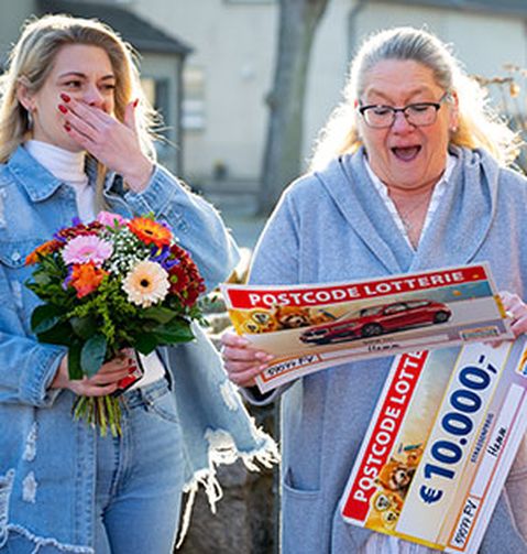 Zwei Frauen reagieren überrascht und freudig. Eine von ihnen hält einen Blumenstrauß, die andere einen großen Lotterie-Scheck über 10.000 €.