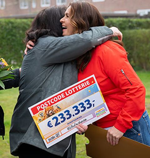 Zwei Frauen umarmen sich freudig und halten einen großen Lotterie-Scheck über 233.333 € in den Händen. Eine Person neben ihnen trägt einen Blumenstrauß.