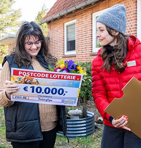 Zwei Frauen stehen vor einem Haus. Eine hält einen großen Umschlag, die andere ein Schild mit der Aufschrift „10.000 €“ und „Postcode“.