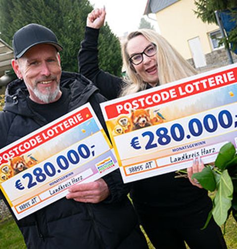 Zwei Personen lächeln und halten große Lotterie-Schecks über jeweils 280.000 € in den Händen. Sie stehen im Freien, im Hintergrund sind Bäume und ein Haus zu sehen.