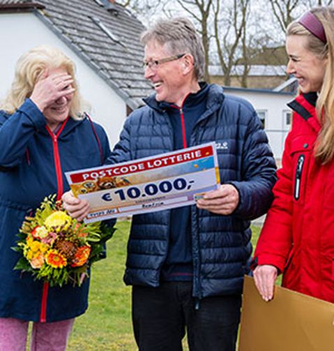 Drei lächelnde Personen stehen im Freien und halten einen großen Scheck über 10.000 € von der Postcode Lotterie. Eine Frau trägt einen Blumenstrauß.