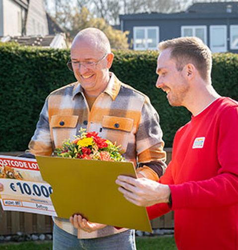 Zwei Männer lächeln im Freien. Einer von ihnen hält einen Blumenstrauß und einen großen Lotterie-Scheck über 10.000 €. Im Hintergrund sind Häuser und viel Grün zu sehen.
