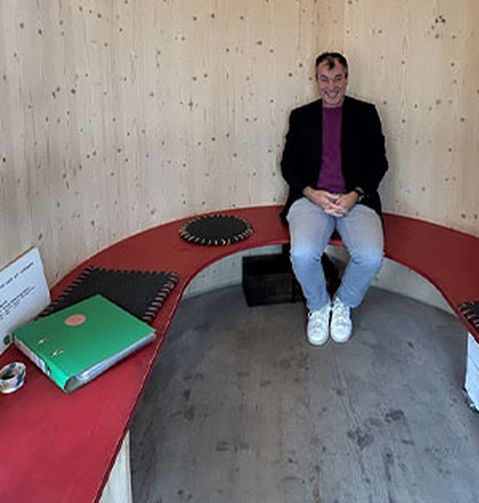 Eine Person sitzt auf einer geschwungenen roten Bank in einem Holzraum. In der Nähe liegen ein grüner Ordner und ein kleines Behältnis.