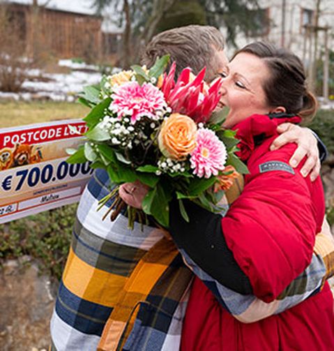 Zwei Personen umarmen sich freudig, eine von ihnen hält einen Blumenstrauß. Im Hintergrund ist ein großer Scheck über 700.000 € zu sehen.