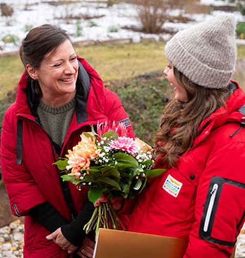 Zwei Personen in roten Jacken lächeln im Freien. Eine von ihnen hält einen Blumenstrauß und einen Umschlag in der Hand. Im Hintergrund sind Schnee und Gras zu sehen.
