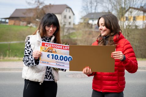 Zwei Frauen stehen im Freien und lächeln. Eine von ihnen hält einen Postcode-Lotterie-Scheck über 10.000 €, die andere ein leeres Schild. Im Hintergrund sind Häuser und Bäume zu sehen.
