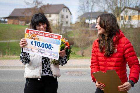 Zwei Frauen stehen im Freien und lächeln. Eine von ihnen hält einen Postcode-Lotterie-Scheck über 10.000 € und einen Blumenstrauß. Im Hintergrund sind Häuser und Bäume zu sehen.