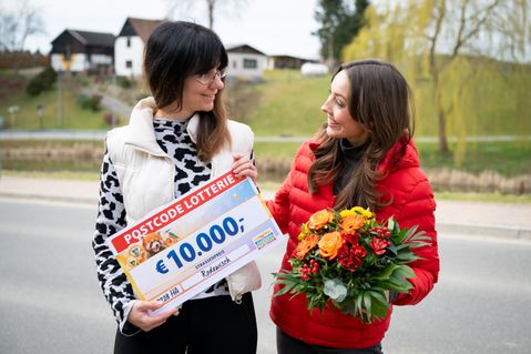 Zwei Frauen lächeln im Freien. Eine von ihnen hält einen großen Lotterie-Scheck über 10.000 €, die andere einen bunten Blumenstrauß.